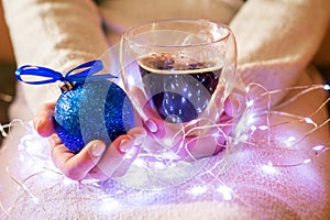 Female hands hold cup of coffee and decorative Christmas ball in Christmas lights