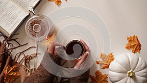 Female hands hold cup of tea,glass teapot,pumpkins,orange maple leaves,twigs,book stand side by side white background,top view,