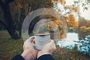 Female hands hold a enameled cup of coffee on autumn landscape.