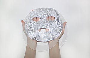 Female hands hold a fabric holographic silver sheet mask for the face.