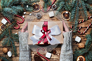 Female hands hold Gift box with bow. Christmas tree and decorations, cane caramel and oranges nuts Wooden background