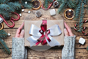 Female hands hold Gift box with bow. Christmas tree and decorations, cane caramel and oranges nuts Wooden background