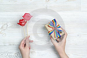 Female hands hold a gift box with rainbow LGBT ribbon nd pair of decorative lips on a light wooden table
