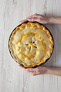Female hands hold homemade apple pie over white wooden background, top view. Flat lay, overhead, from above