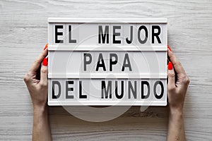 Female hands hold lightbox with `El Mejor Papa Del Mundo` words over white wooden background. Overhead, from above, flat lay. photo