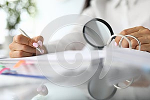 Female hands hold magnifying glass and pens next to documents.