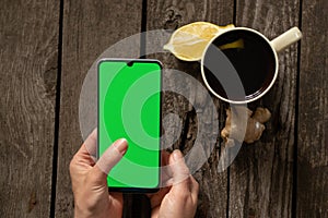 Female hands hold a mobile phone and a cup of tea with lemon on an old wooden table