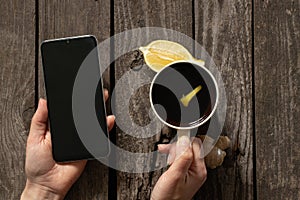 Female hands hold a mobile phone and a cup of tea with lemon on an old wooden table