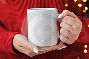 Female hands hold a mockup white tea mug in a red winter sweater, gold bokeh