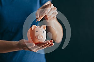 Female hands hold a pink piggy bank and puts a coin there. The concept of saving money or savings, investment