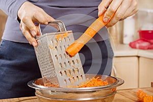 Female hands hold a raw carrot and grind it into small pieces on a metal kitchen grater.