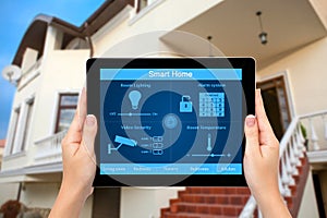 Female hands hold a tablet with system smart house on the background of the house