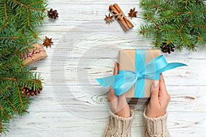 Female hands hold wrapping christmas gift box with blue ribbon on white wooden table. Top view