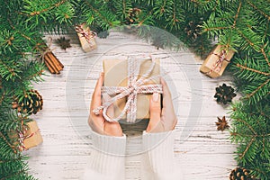 Female hands hold wrapping christmas gift box with pink ribbon on white wooden table. Top view. Toned