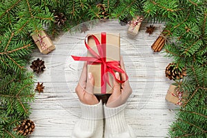 Female hands hold wrapping christmas gift box with red ribbon on white wooden table. Top view