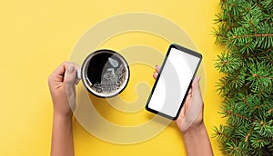 Female hands holding black mobile phone with blank white screen and mug of coffee. Mockup image with copy space. Top view banner