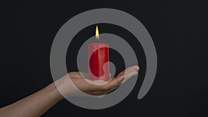 Female hands holding a burning red candle on dark background