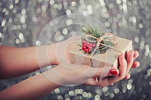 Female hands holding Christmas gift box with branch of fir tree, shiny xmas background. Holiday gift and decoration. Toning.