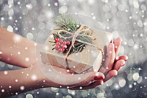 Female hands holding Christmas gift box with branch of fir tree, shiny xmas background. Holiday gift and decoration. Toning.
