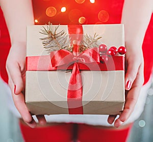 Female hands holding Christmas gift box with red ribbon, branch of fir tree on shiny xmas background