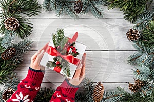 Female hands holding Christmas gift box on white wooden background with Fir branches, pine cones.