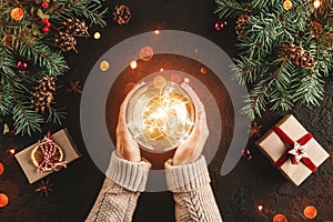 Female hands holding Christmas glowing ball on holiday background with Fir branches, gifts.