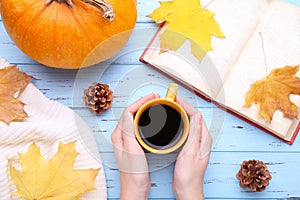 Female hands holding cup of black coffee with autumn leaves, book and pumpkin on blue