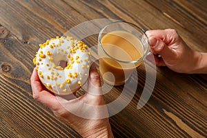 Female hands holding cup of coffee and donut in a white glaze with yellow confectionery sprinkles on a wooden background