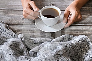 Female hands holding a cup of hot beverage, tea or coffee on a wooden table background with a cozy knitted warm winter