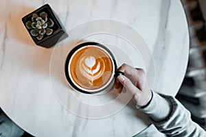 Female hands holding a cup of latte on the background of the table in a cafe. A lover of hot coffee in the morning enjoys