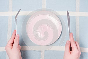 Female hands holding a fork and and knife next to an empty pink plate on a blue tablecloth background close-up top view.