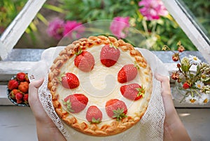 Female hands holding a homemade strawberry cake near window on summer garden background