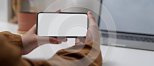 Female hands holding horizontal mock up smartphone while sitting at worktable with mock up laptop