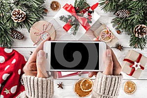 Female hands holding mobile phone. Christmas gift boxes with red decoration, Fir branches, pine cones, paper card note on holiday