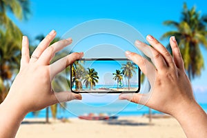 Female hands holding mobile smart phone and making photo of amazing beach with palms and ocean. Vacation and relaxation concept