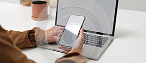 Female hands holding mock up smartphone while sitting at worktable with mock up laptop in home office
