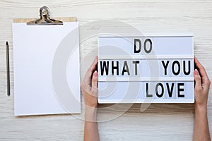 Female hands holding modern board with text `Do what you love`. Blank noticepad with pencil. White wooden background, top view.