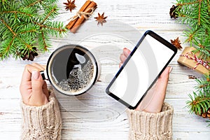 Female hands holding modern smartphone with mosk up and mug of coffee on wooden table with christmas decoration. top