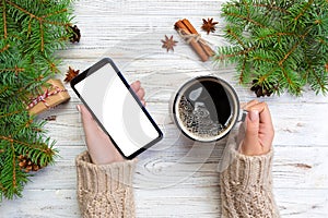 Female hands holding modern smartphone with mosk up and mug of coffee on wooden table with christmas decoration. top view