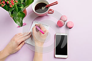 The female hands holding pen. The phone and french macarons on trendy pink desk.