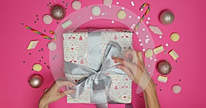 Female hands holding present box wrapped in festive paper and tied with backers twine on pink background with decor