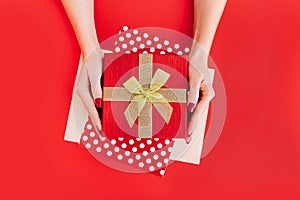 Female hands holding red goft box present with golden bow on red background. Festive backdrop for holidays: Birthday