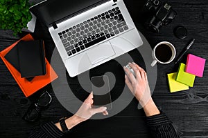 Female hands holding a smart and typing on the keyboard of a laptop on black wooden table.