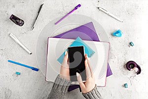 Female hands holding smartphone at workplace with notepads, pens and other office supplies. Women`s business concept