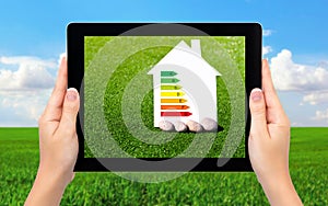 female hands holding a tablet with a house and sign energy saving on a background of field