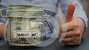 Female hands holding thumb up like sign Glass jar full of American currency dollars cash banknote with text SWEAR JAR