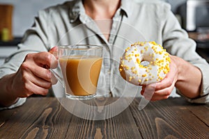 Female hands holding transparent cup of coffee and donut in a white glaze with yellow confectionery sprinkles on a kitchen