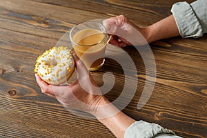 Female hands holding cup of coffee and donut in a white glaze with yellow confectionery sprinkles on a wooden background