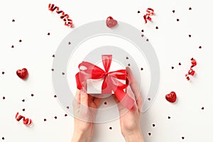 Female hands holding white gift box with red ribbon bow over white background with party streamers, confetti and candy. Happy