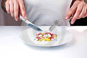 Female hands holds knife and fork and in front of her lie multicolored pills on a white plate. Taking vitamins and supplements.